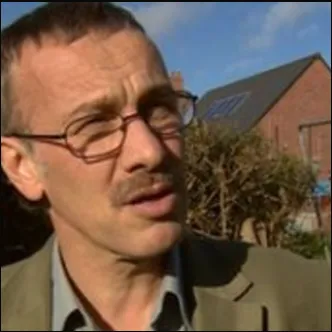 A man with glasses and a mustache is seen outdoors, standing in front of buildings with a blue sky background.