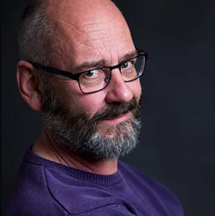 A man with a bald head, grey beard, and eyeglasses is looking over his shoulder, wearing a purple shirt against a dark background.