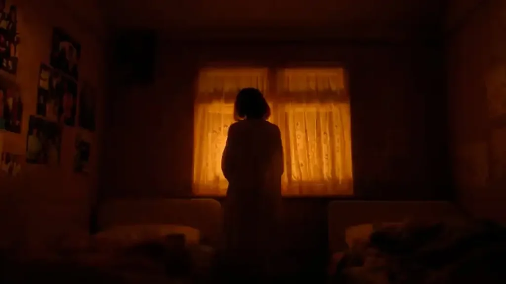 A person in a dimly lit room stands facing a window with orange-hued curtains.