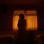 A person in a dimly lit room stands facing a window with orange-hued curtains.