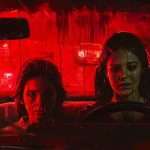 Two individuals sit in a car at night with a red, ominous light illuminating their faces. Their expressions are serious and contemplative as they look out the windshield.