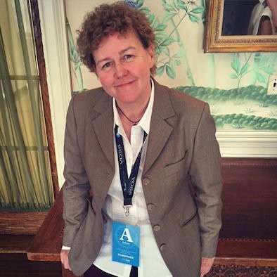 Person with curly hair in a taupe blazer and white shirt stands indoors, wearing a lanyard with a blue "A" badge. They smile slightly at the camera.