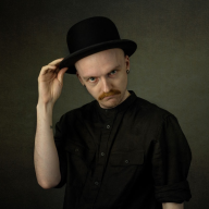 A person with a mustache, wearing all black and a black bowler hat, lifts their hat slightly while looking directly at the camera.