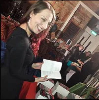 A person wearing a black top is smiling while holding an open book. Other people are visible in the background in a room with brick walls and various items on tables.