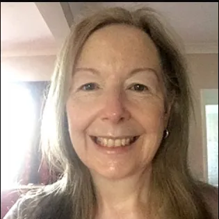 A woman with long blonde hair is smiling directly at the camera. She is indoors with light coming from the left side of the image.