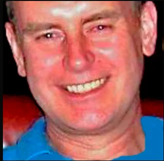 A smiling man with short hair wearing a blue shirt. The background is dark and not clearly visible.