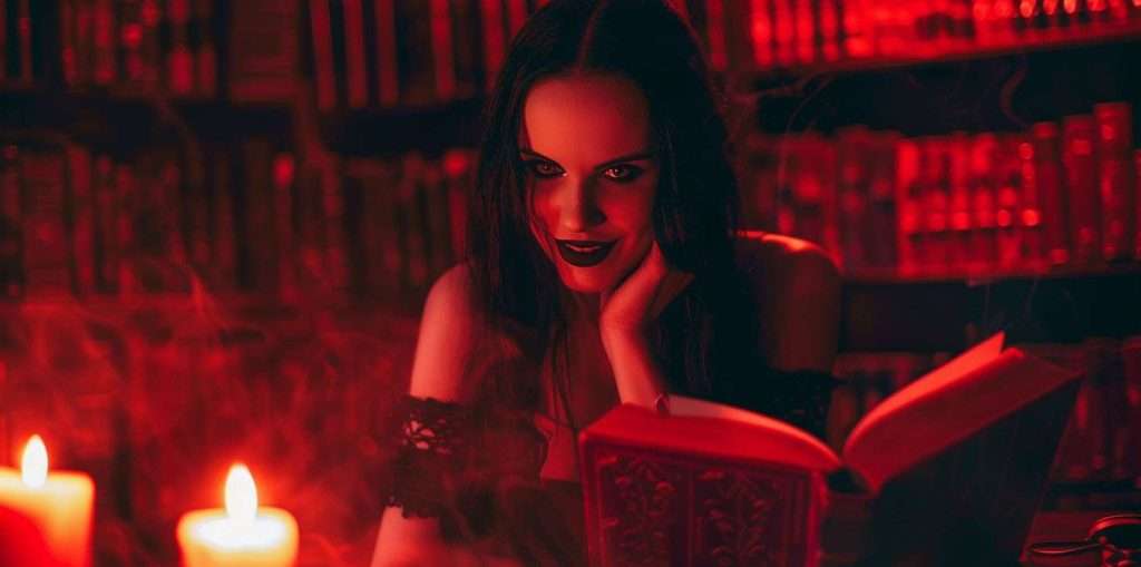 A woman with dark hair and gothic makeup reads a book in a dimly lit room with red lighting and candlelight.