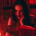 A woman with dark hair and gothic makeup reads a book in a dimly lit room with red lighting and candlelight.