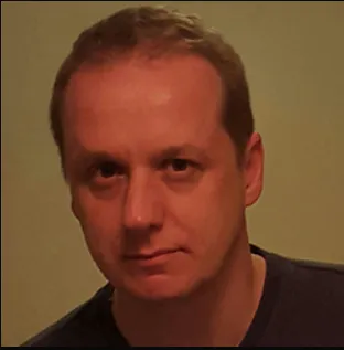 A man with short hair, wearing a dark shirt, looks directly at the camera against a plain background.