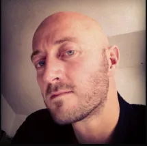 A man with a bald head and a trimmed beard looks at the camera with a neutral expression. He is wearing a black shirt, and the background appears to be indoors.