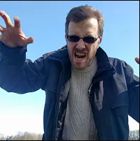 A man wearing sunglasses and a jacket, making a roaring or growling gesture with his hands raised and mouth open, stands under a clear blue sky.