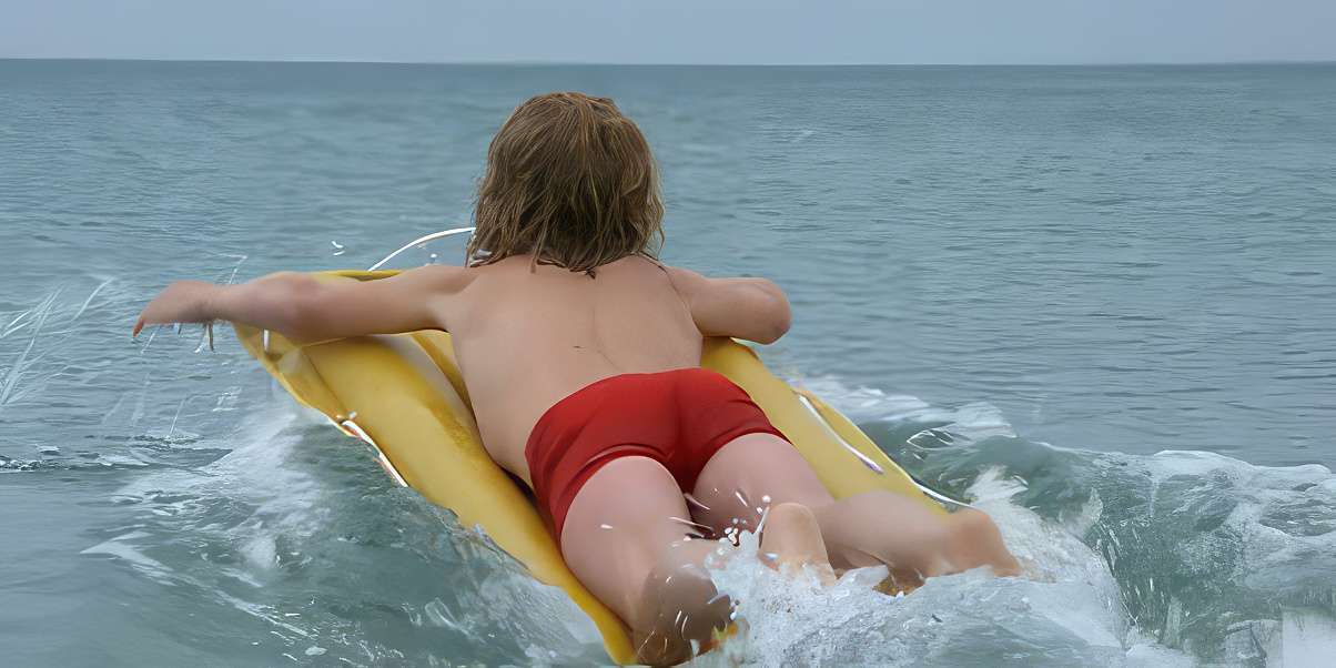 A child in red swim shorts lies on a yellow inflatable mat, floating on the ocean's surface with small waves around.