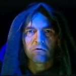 A person with long hair under a dark hood, illuminated by blue light, staring directly at the camera with a serious expression.