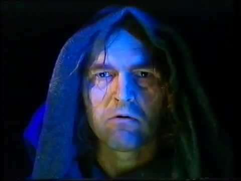 A person with long hair under a dark hood, illuminated by blue light, staring directly at the camera with a serious expression.