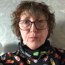A person with curly hair, large glasses, and a serious expression is wearing a colorful top with various patterns.