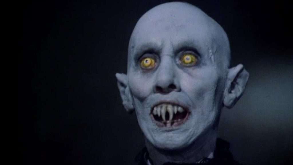 A bald figure with pale blue skin, glowing yellow eyes, pointed ears, and sharp fangs stares ahead with an open mouth, against a dark background.
