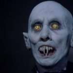 A bald figure with pale blue skin, glowing yellow eyes, pointed ears, and sharp fangs stares ahead with an open mouth, against a dark background.