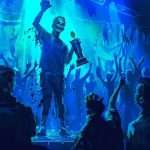 A person holding a trophy stands on a stage under blue lights, surrounded by an exuberant crowd with hands raised in celebration.