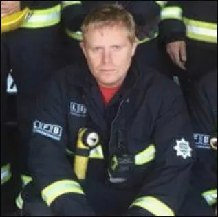 A person wearing a firefighter uniform sits with others in similar attire, holding a helmet. The individual has short blond hair and is looking directly at the camera.
