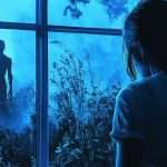 A person stands inside a room in front of a window, looking out at a silhouetted figure with glowing eyes in a mysterious, misty, blue-toned outdoor setting.