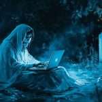A cloaked figure sits among tombstones at night, using a glowing laptop.