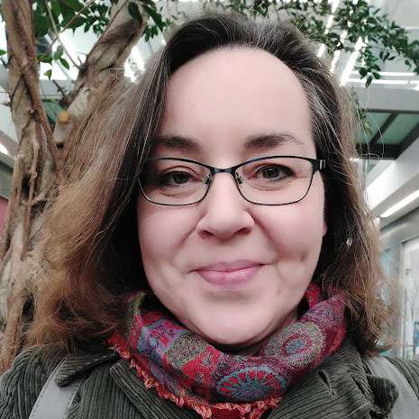 A middle-aged person with glasses and brown hair wearing a patterned scarf smiles at the camera, standing in front of a tree in an indoor setting.