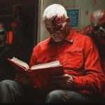 A man with white hair reads a book on a train, surrounded by zombies. He is seated and appears calm despite the gruesome figures near him.