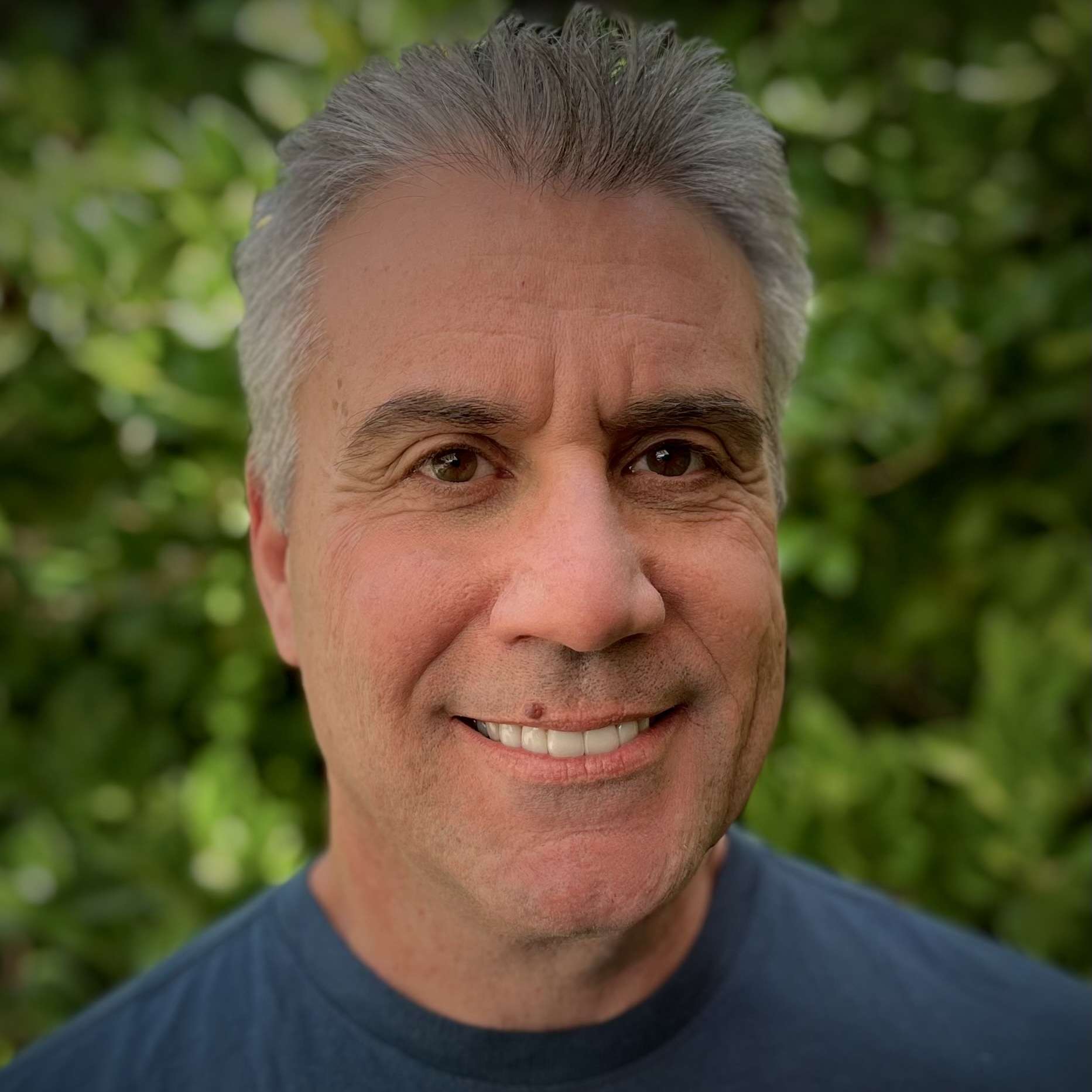 A person with short gray hair, wearing a dark blue shirt, smiles in front of a leafy green background.