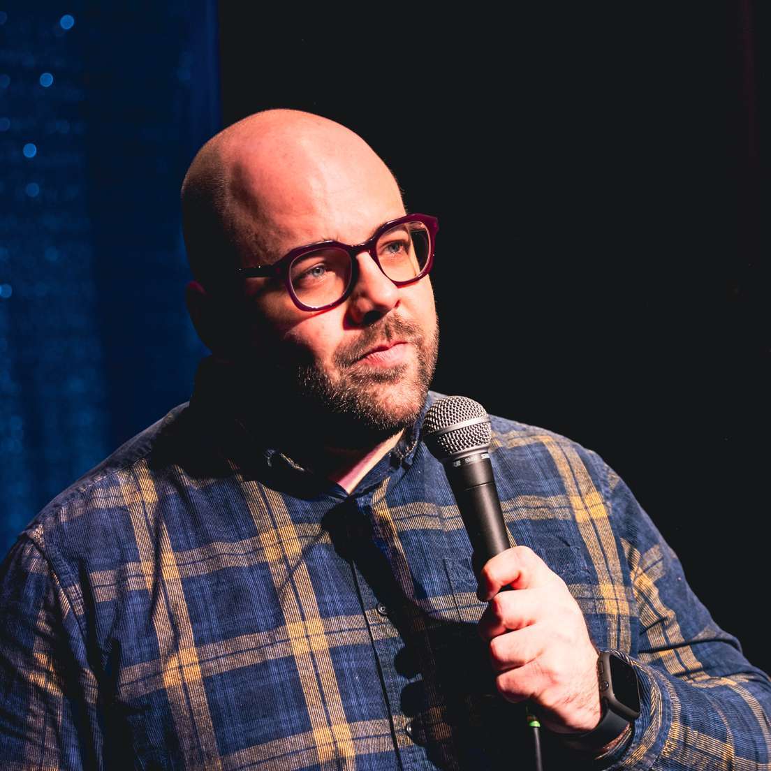A person with glasses and a plaid shirt is holding a microphone on stage.