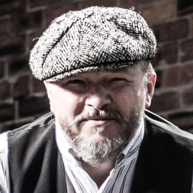 A man with a beard wearing a flat cap and a vest stands against a brick wall.