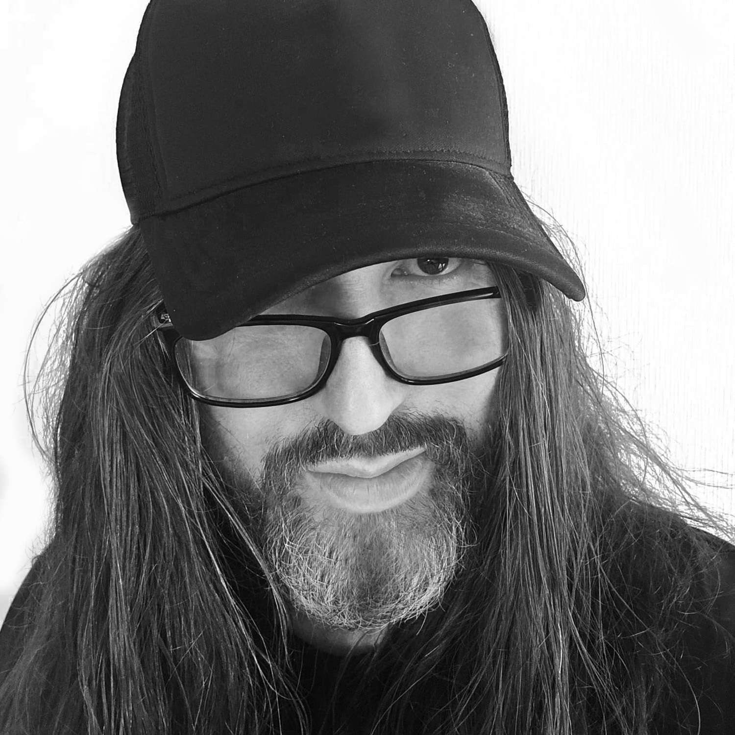 Person with long hair, beard, and glasses wearing a baseball cap, looking at the camera. Black and white image.
