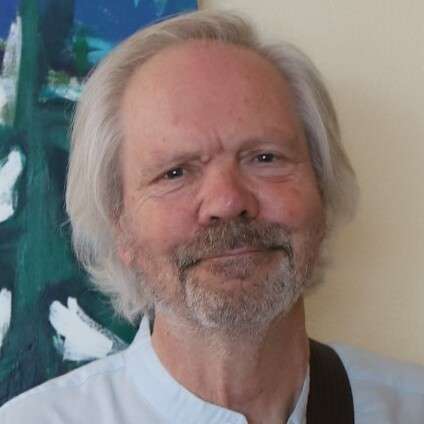 An older man with white hair and a beard wearing a light-colored shirt stands in front of an abstract painting.