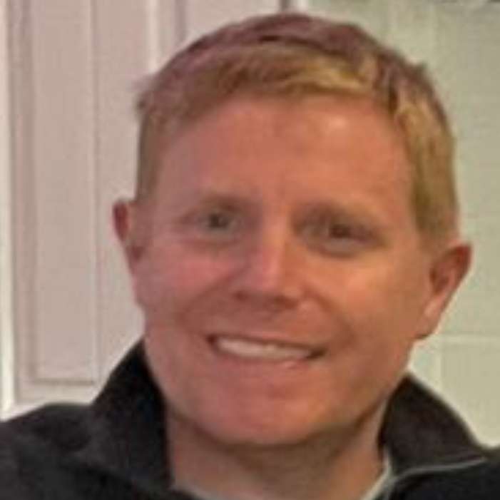 A person with short light hair wearing a dark jacket, smiling at the camera indoors.