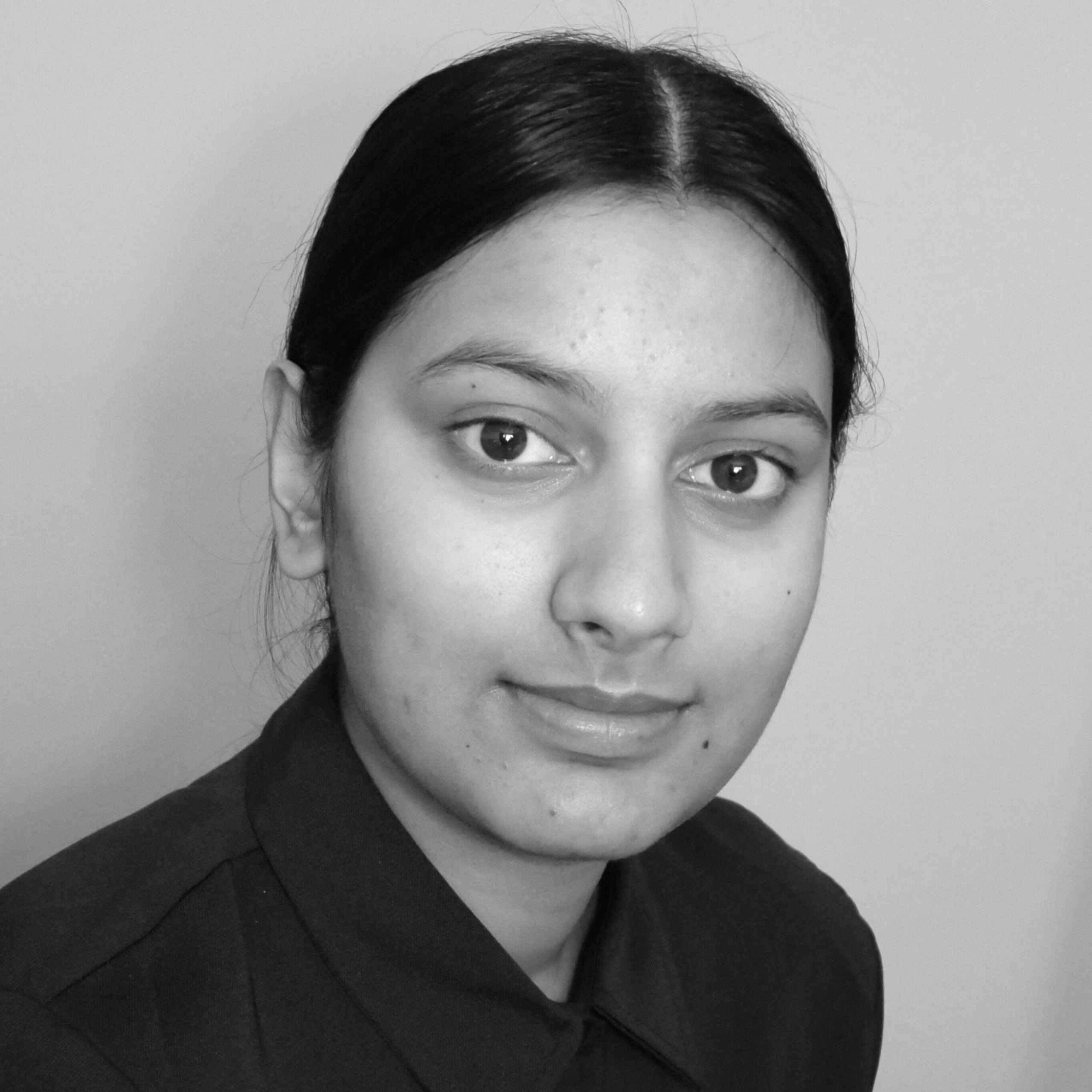 A person with dark hair pulled back, wearing a collared shirt, looks straight at the camera. The image is in black and white.