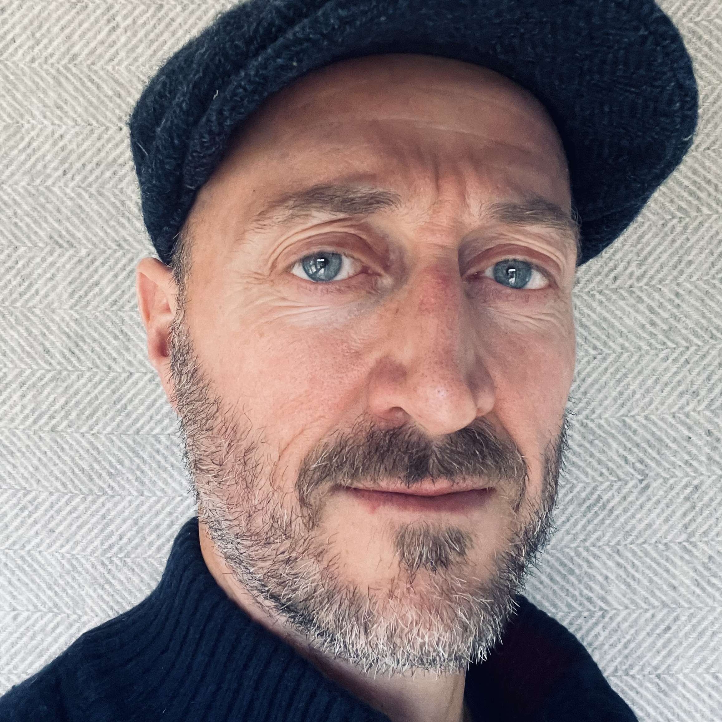 Man wearing a black cap and sweater, looking directly at the camera, with a neutral expression.