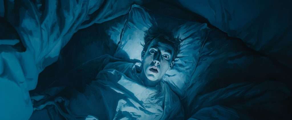 A person lies in bed under a blanket, staring upward with wide eyes and a startled expression, illuminated by bluish light in a dark room.