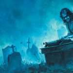 A woman uses a typewriter on a desk in a graveyard at night, surrounded by gravestones, under blue, eerie lighting.