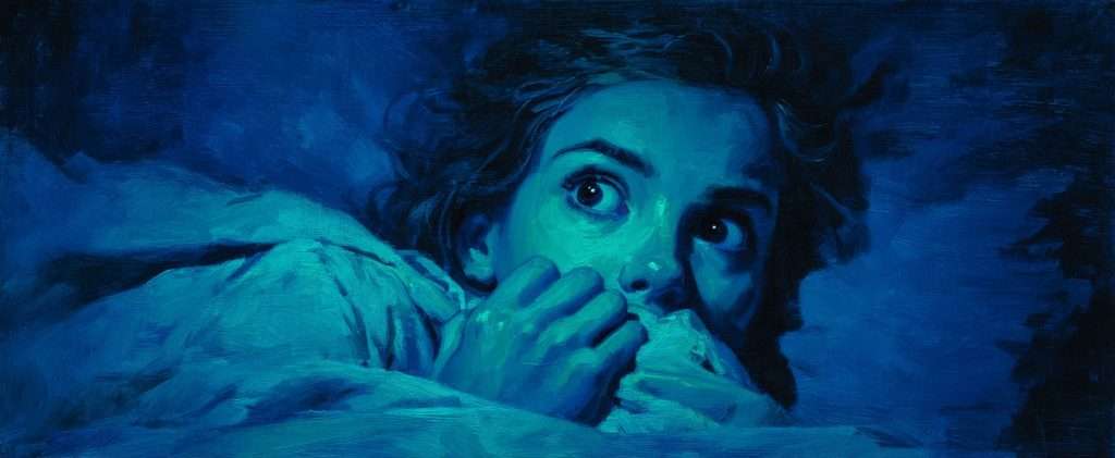 A person lying in bed, illuminated by blue light, looks wide-eyed and fearful while clutching a blanket close to their face.