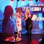 Two women stand on stage at an award event; one holds a certificate and microphone, while the other wears a white dress with red handprints. The screen behind displays "Modern Gothic Award.