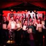 A group of people stand on stage holding certificates, some raising their fists and smiling, in front of a red-lit background that partially reads "WINNERS.
